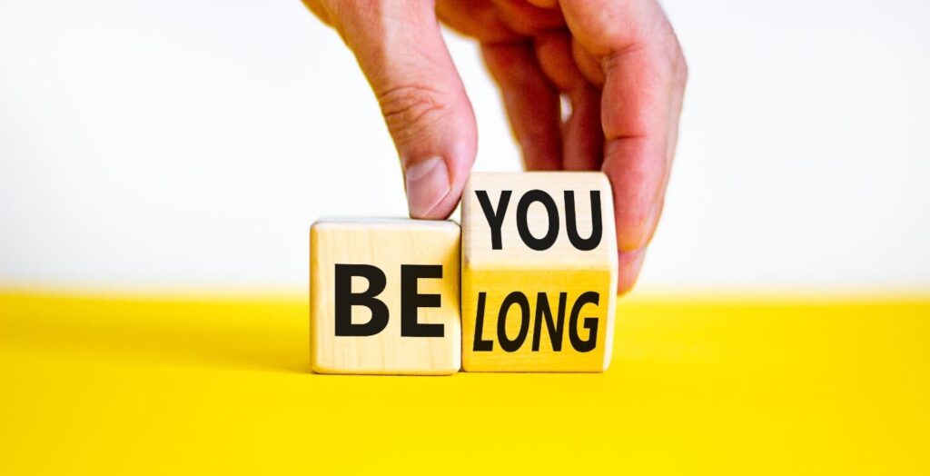 you belong in Black letters on a light brown blocks on a yellow table with white background. A hand grabs one of the blocks
