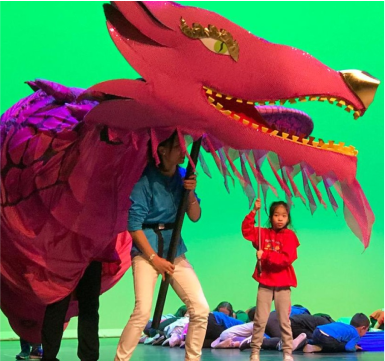 man holds a big red dragon. A little girl points at dragon. children lie down in the background near a green coloured screen