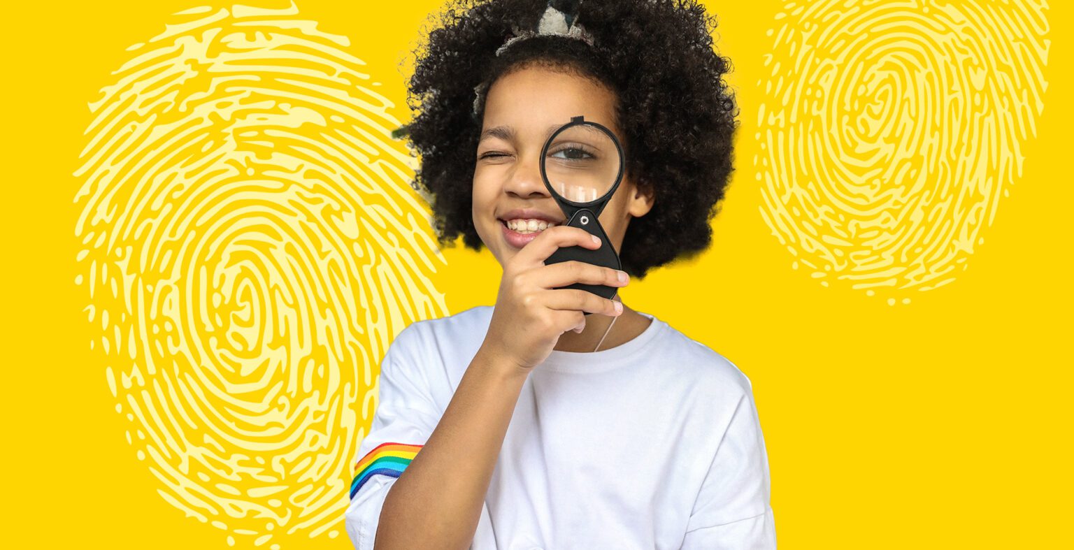 Little girl in shite shirt and rainbow sleeve holds a magnifying glass to her eye and smiles at camera. Yellow background with white fingerprint marks.